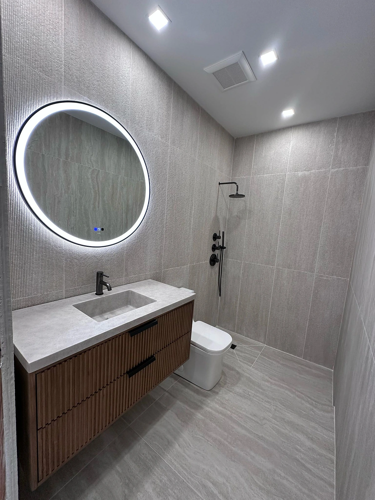 Grey tile bathroom with round backlit mirror and floating wood vanity